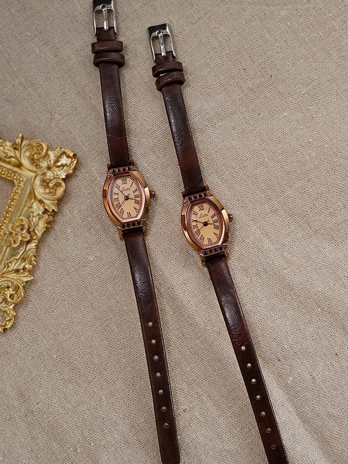 Un par de relojes de cinturón de cuero marrón retro con detalles dorados y pedrería sobre tela beige.