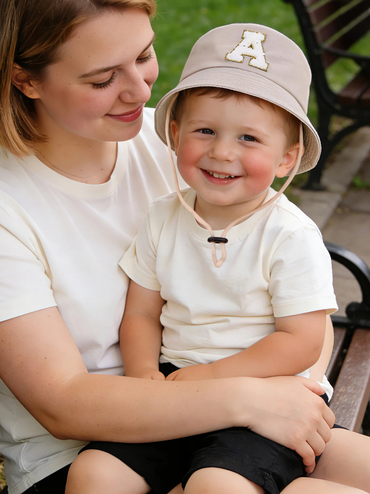 A woman is holding a young boy wearing a beige letter print sun hat.