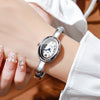 A close-up of a silver quartz watch bracelet worn on a wrist, with a hand displaying manicured nails.