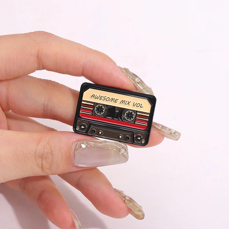 A hand holds a black cassette enamel pin brooch with the text AWESOME MIX VOL visible on the label.