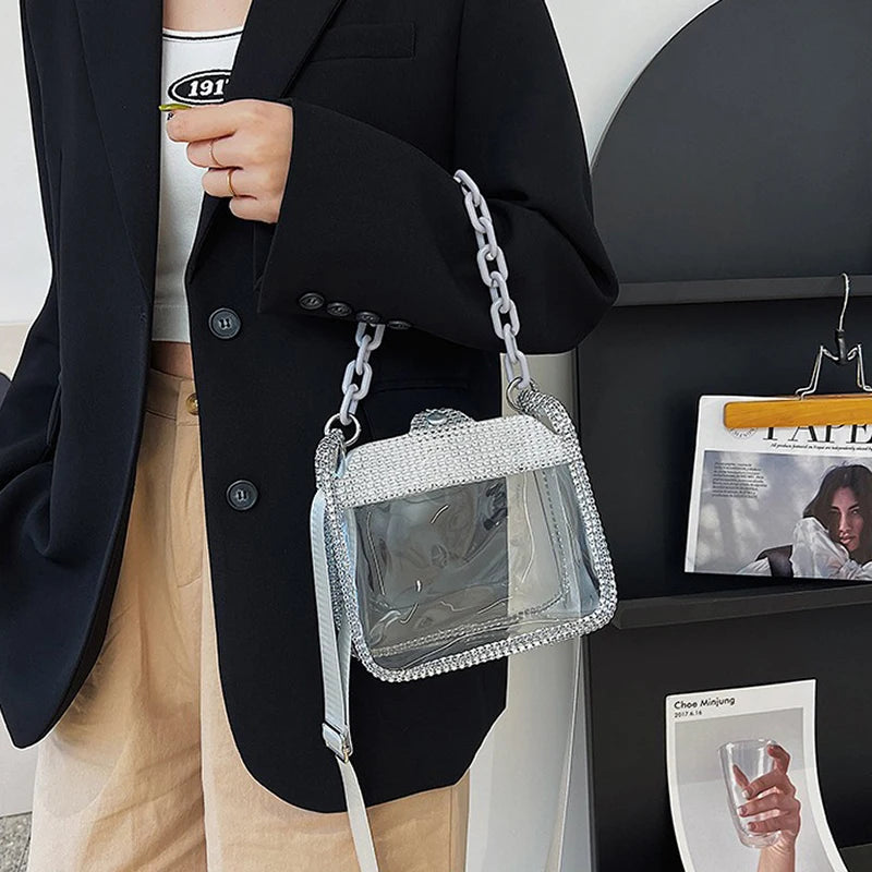 Una persona vistiendo un blazer negro y pantalones color arena sostiene un bolso cruz transparente con cadena de plata.