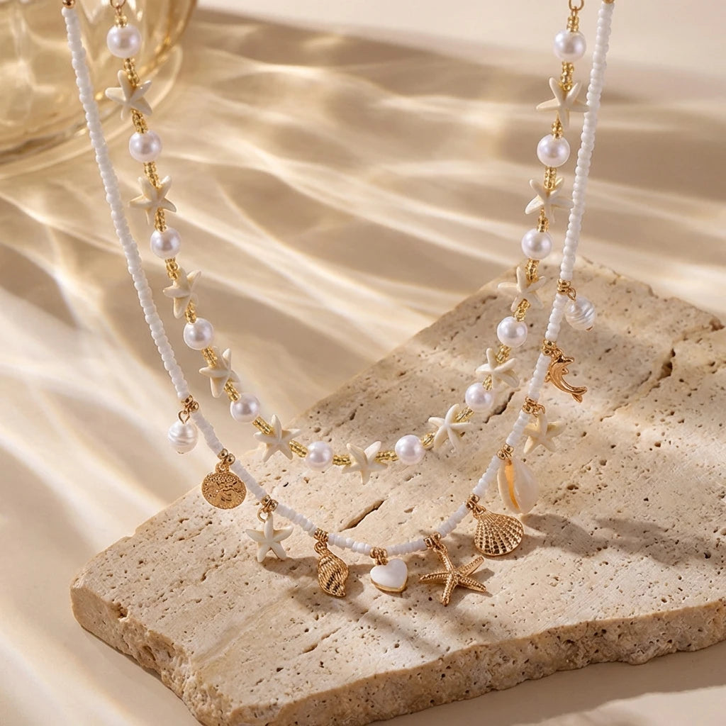 A delicate gold and white beaded necklace featuring starfish and shell charms displayed on a light stone.