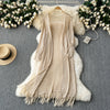 A beige knitted shawl cardigan draped over a white fluffy rug, surrounded by white flowers and a straw hat.