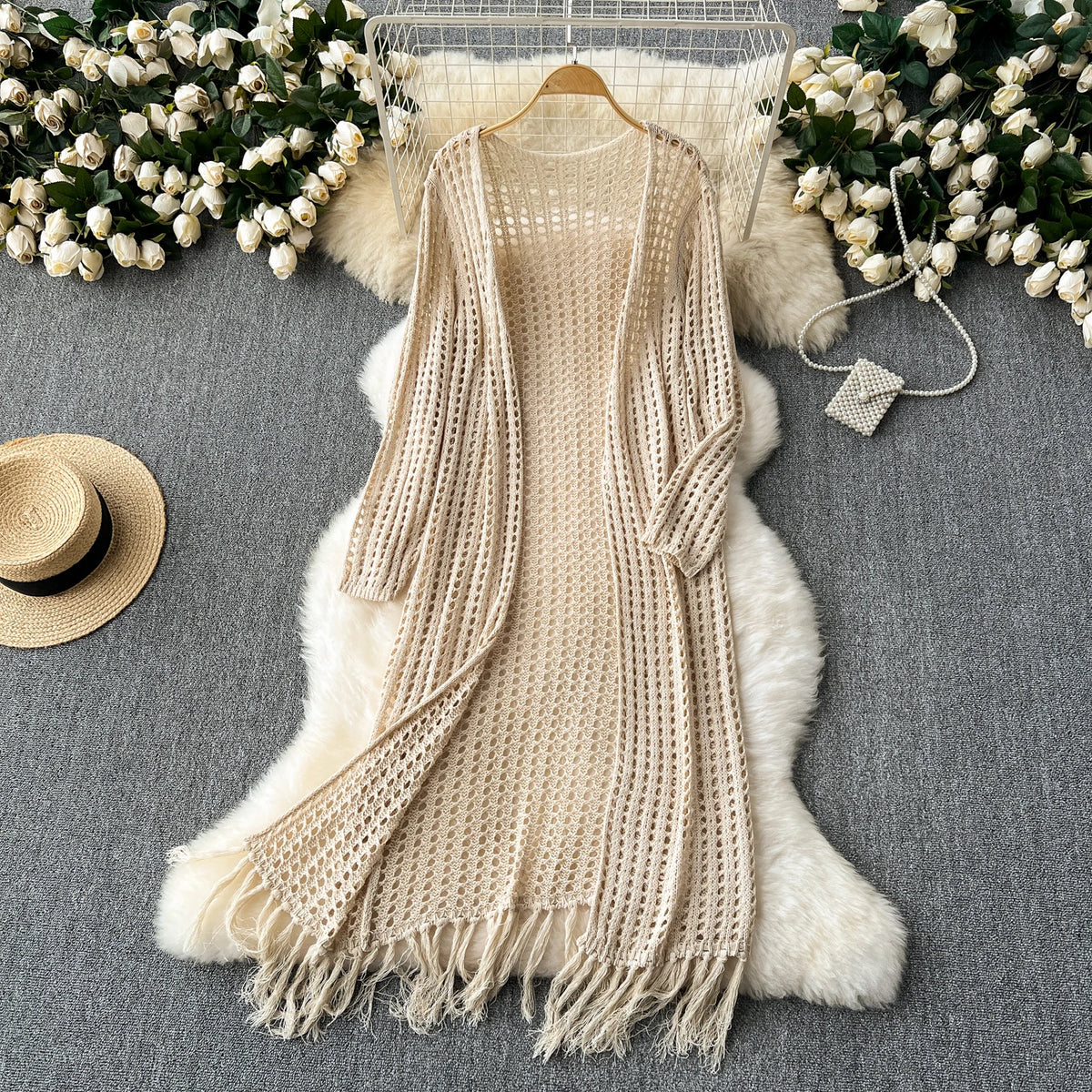 A beige knitted shawl cardigan draped over a white fluffy rug, surrounded by white flowers and a straw hat.