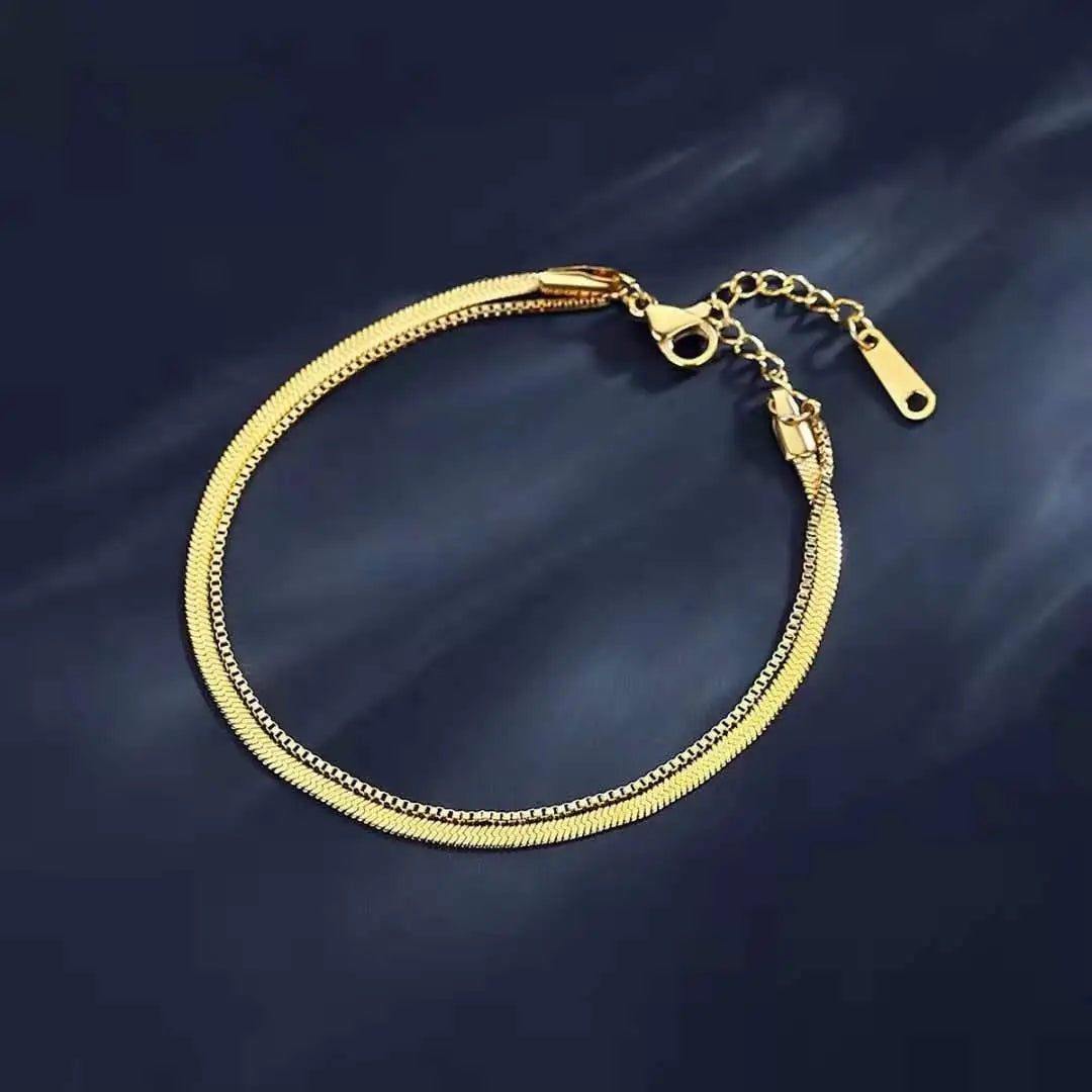 A gold double snake anklet with a chain and clasp is displayed against a dark background.