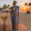 A plus size model wearing a blue and orange African silk printed maxi dress on a wooden deck near a tropical beach at sunset.