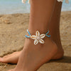 A woman wearing a white flower anklet with blue beads and cowrie shells on her foot on a sandy beach.