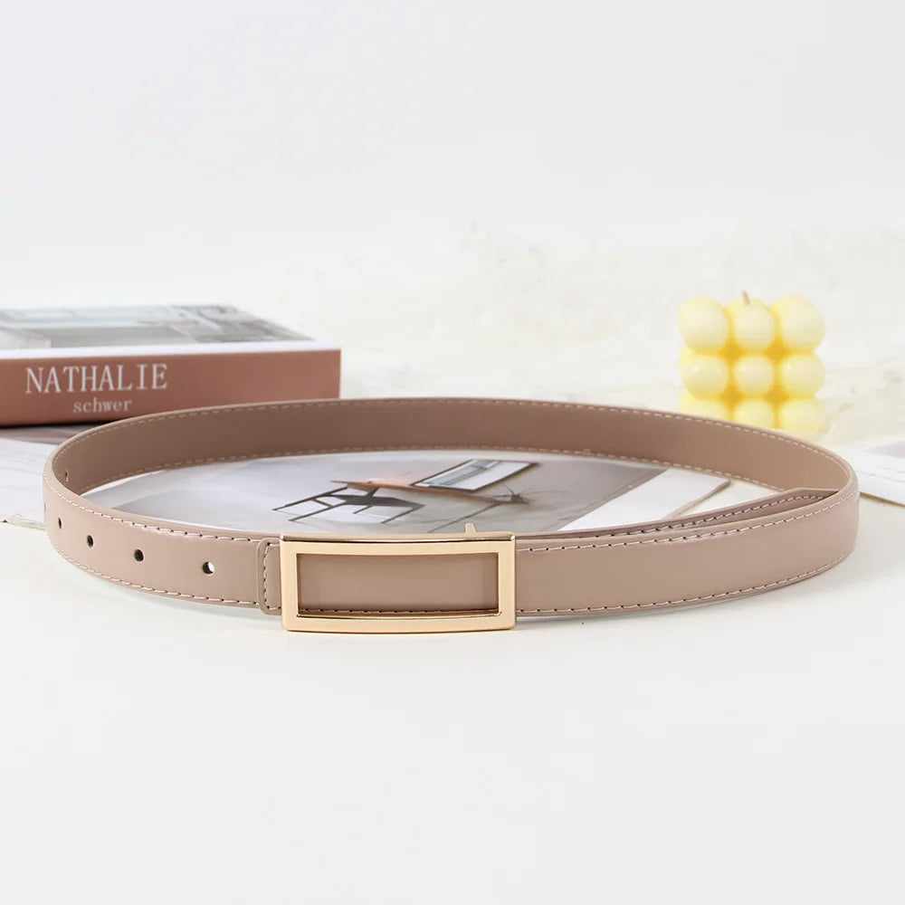 A beige faux leather belt with a gold buckle is displayed next to a book and some yellow corn cobs.