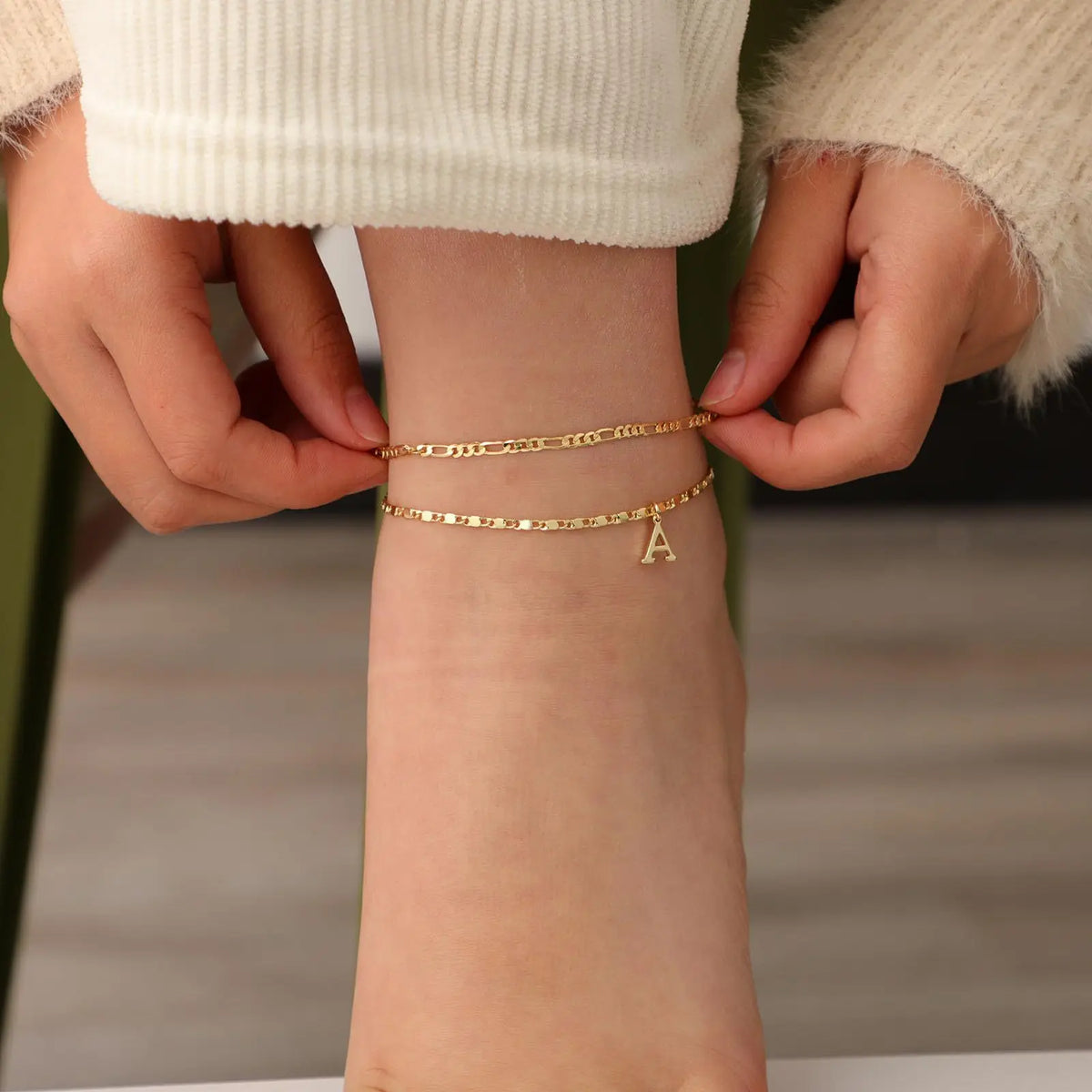 A close-up of a woman's wrist wearing layered gold letter anklets.