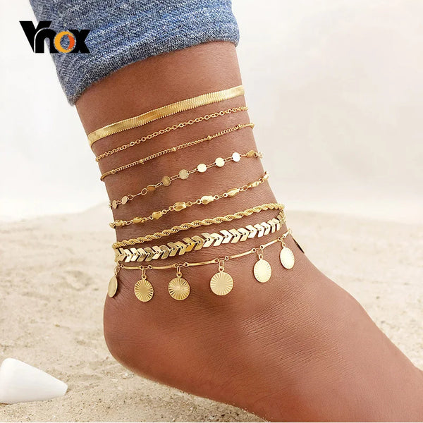 A close-up of multiple layered gold stainless steel anklets worn on a person's foot.