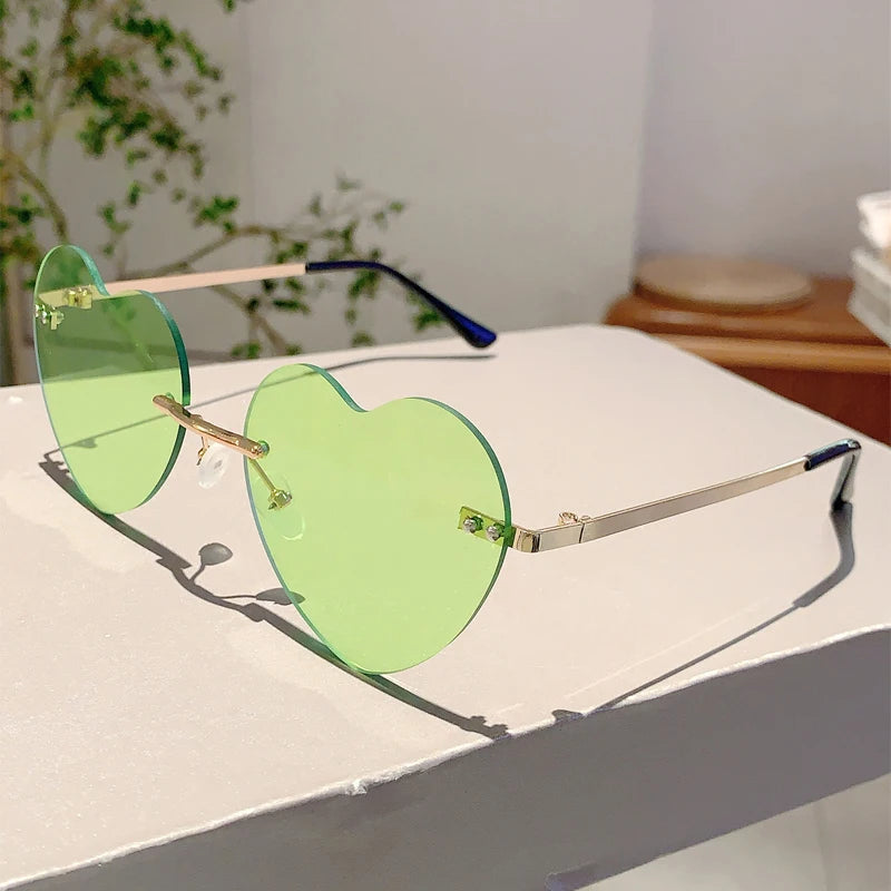 Vintage Herzform-Sonnenbrille ohne Rand mit grün getönten Linsen und goldenen Akzenten auf weißem Untergrund.
