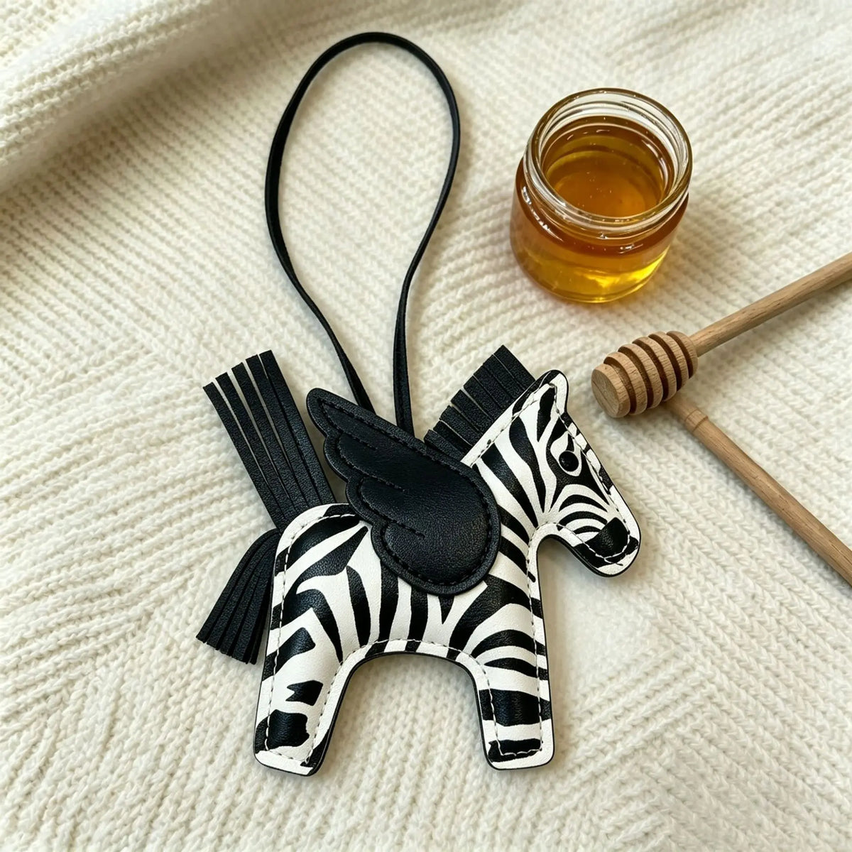 A black and white zebra-print horse charm pendant is displayed next to a small glass jar of honey on a cream knitted fabric.