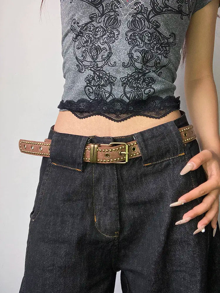 A close-up of dark denim pants and a black lace top featuring a brown belt with bronze rivets.