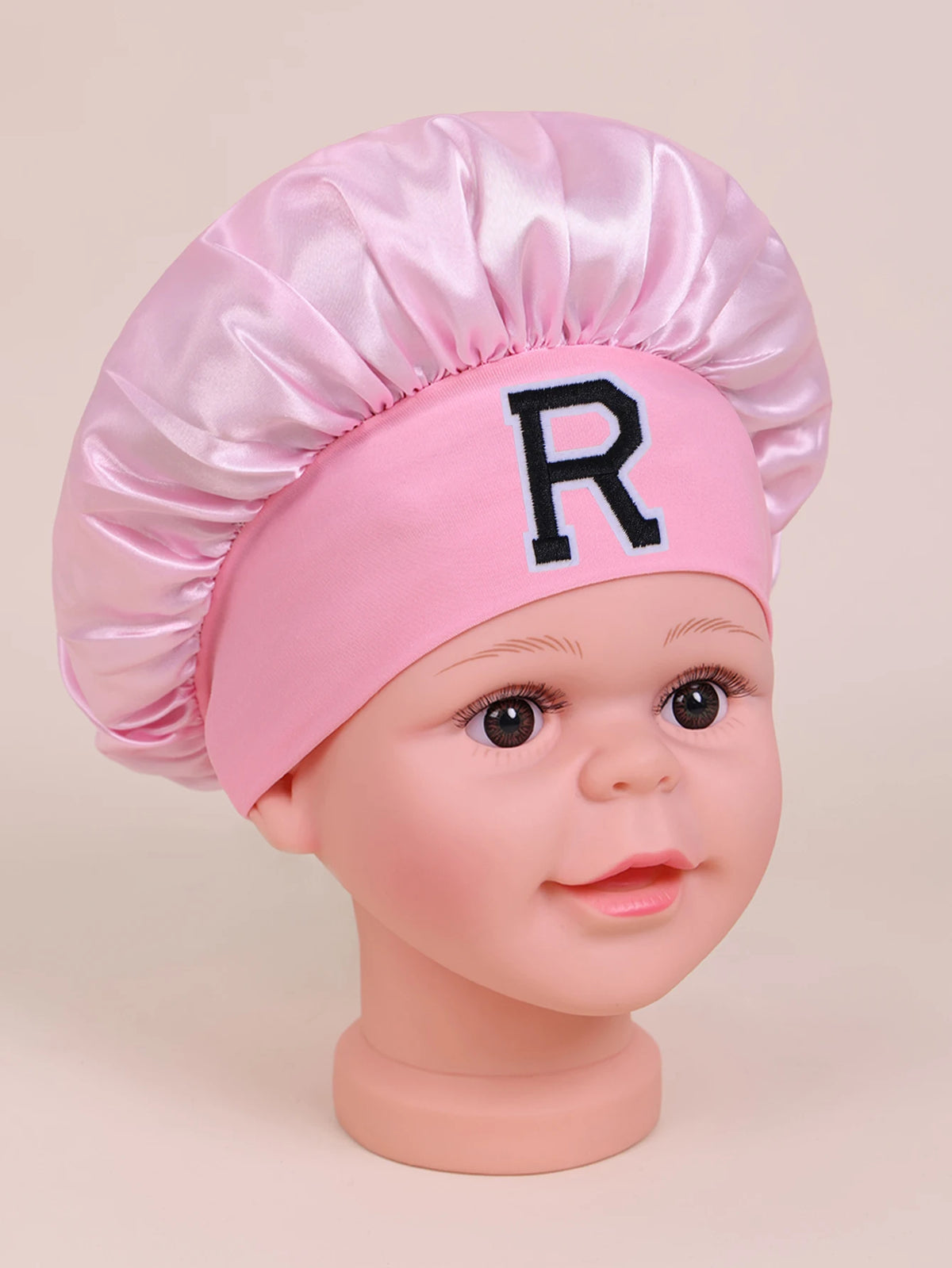 Pink satin sleep cap with a ruffled top and a black letter R on a baby doll head.