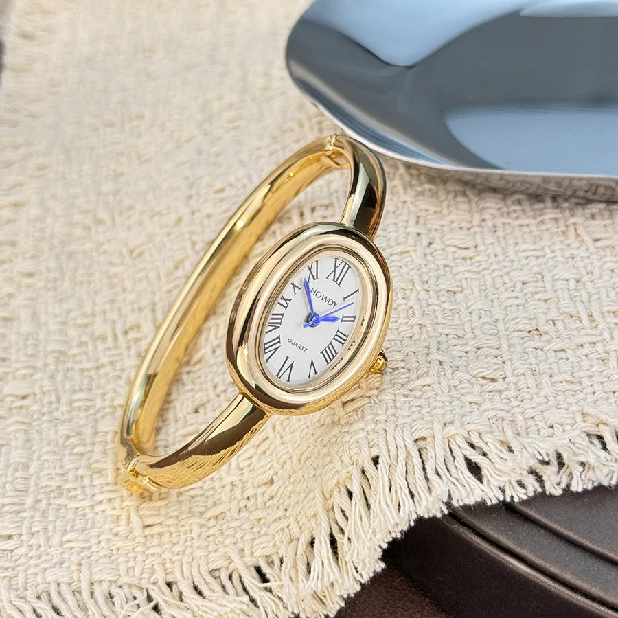 A gold-toned quartz watch with a white face and blue hands is displayed on a beige linen fabric next to a silver dish.
