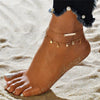 A close-up of a woman's foot wearing an elegant geometric anklet bracelet with gold chains and small charms on a sandy beach.