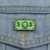 A green enamel dollar banknote brooch featuring an ocean shell charm pinned to blue denim fabric.