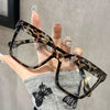 Una mano sostiene unas gafas cuadradas estampadas con patrón de tortuga contra un fondo gris.