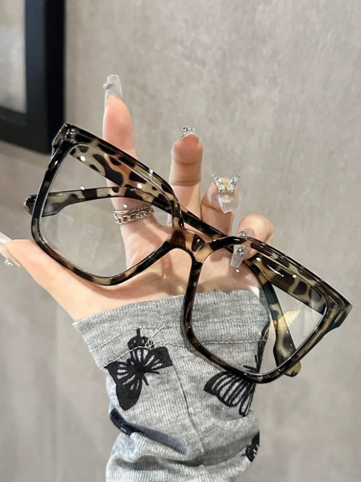 Una mano sostiene unas gafas cuadradas estampadas con patrón de tortuga contra un fondo gris.