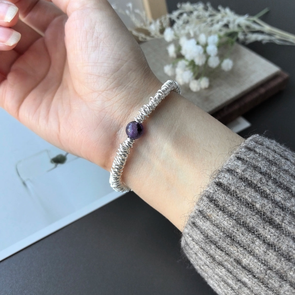 A hand wearing a sterling silver double heart charm bracelet with a purple gemstone, resting on a wooden surface.