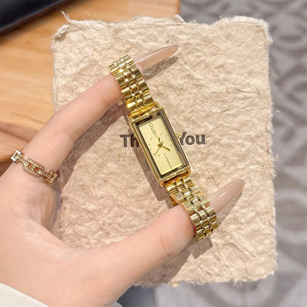 A hand holds a vintage gold stainless steel quartz watch on a beige textured surface.