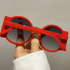 A hand holds a pair of bright red round sunglasses with dark lenses against a light gray background.