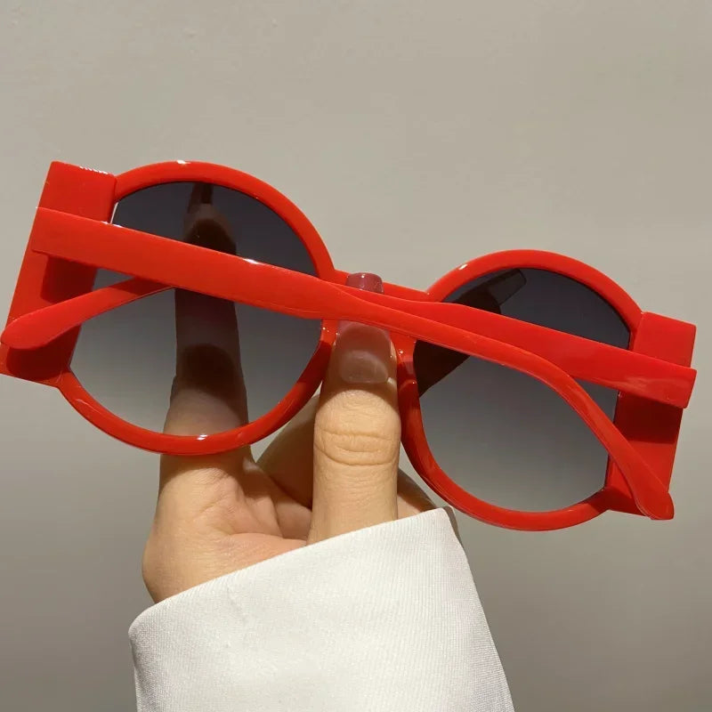 A hand holds a pair of bright red round sunglasses with dark lenses against a light gray background.