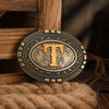 A vintage western belt buckle with a gold-toned 'T' design set in an antique brass frame, resting on burlap.