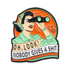An enamel pin featuring a stylized face looking through a telescope with the text OH LOOK nobody gives a shit