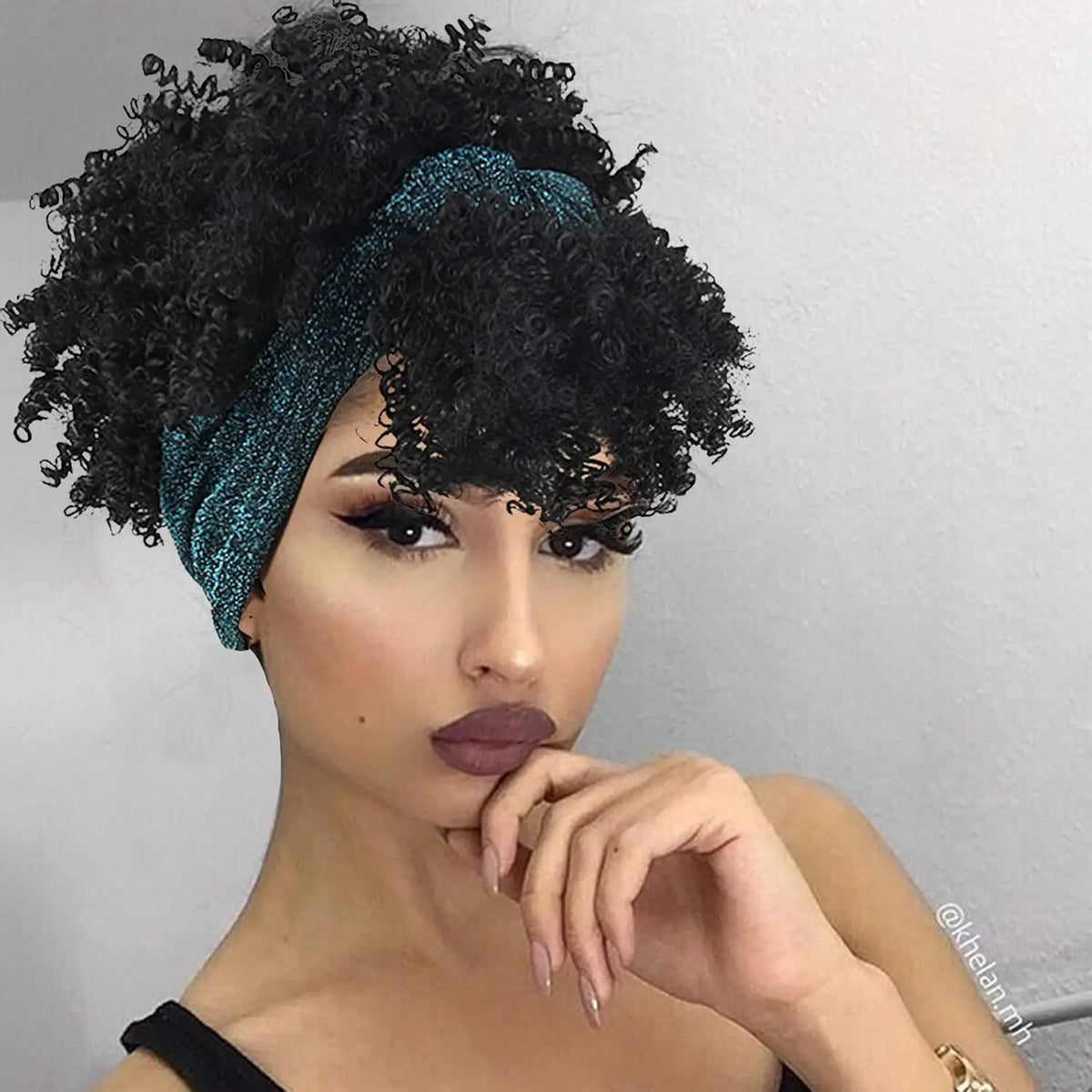 Modelo vistiendo una peluca afro rizada oscura con cinta azul verdosa y maquillaje dramático.