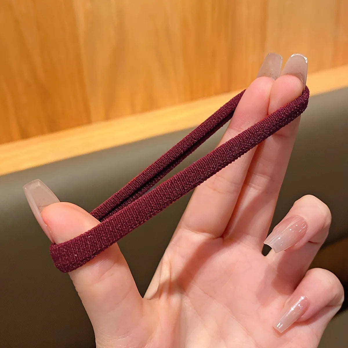 A hand holds a dark maroon elastic hair tie against a blurred wooden background.
