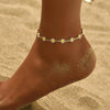 A close-up of a foot wearing a delicate yellow enamel daisy chain anklet against a sandy background.