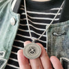Collar de plata con dije del logo de espada Berserk de anime, sostenido contra una camisa a rayas oscuras y una chaqueta verd