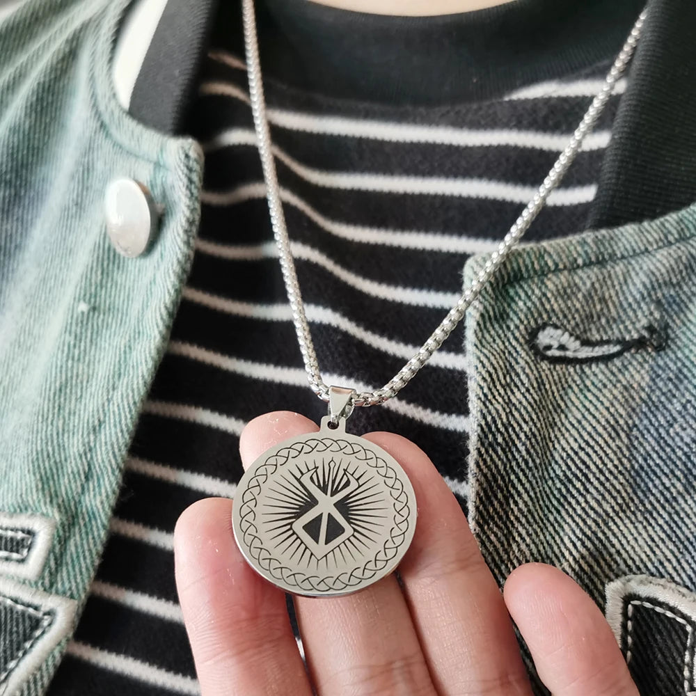 Collar de plata con dije del logo de espada Berserk de anime, sostenido contra una camisa a rayas oscuras y una chaqueta verd