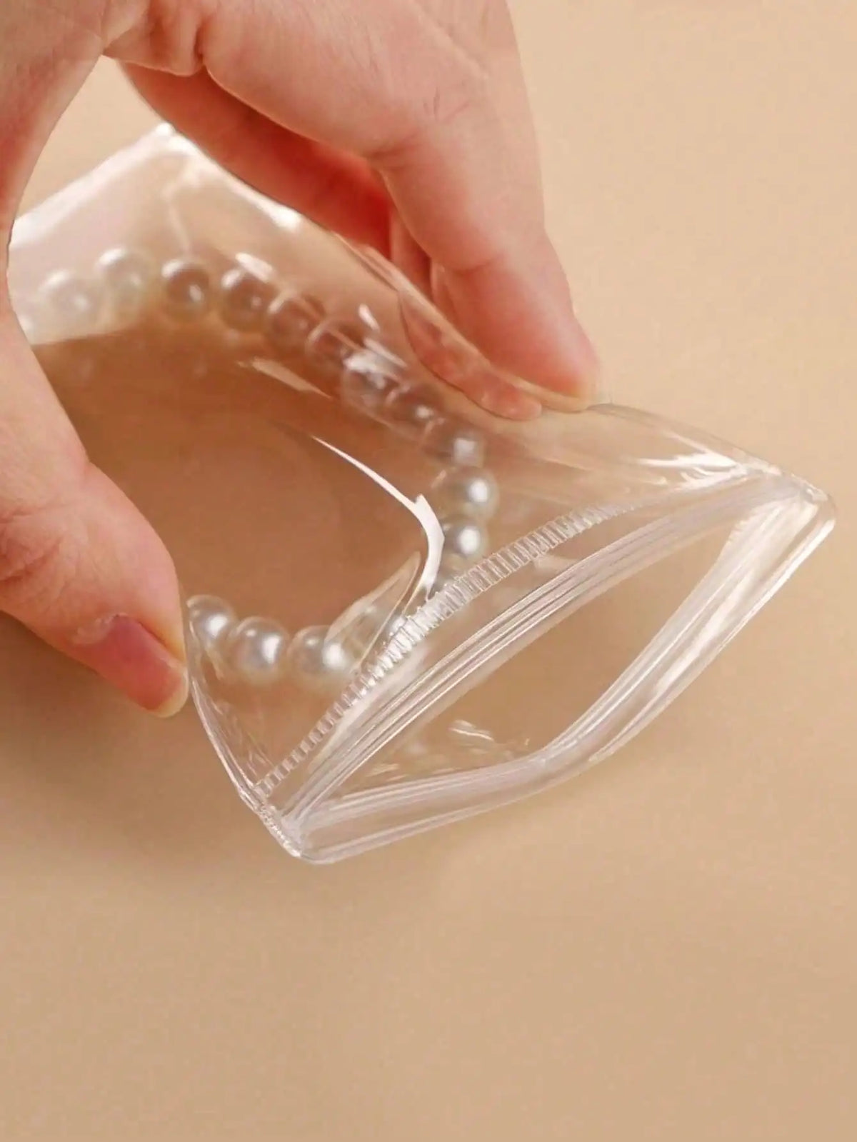 A hand is holding a clear self-sealing jewelry storage pouch filled with pearl-like beads.