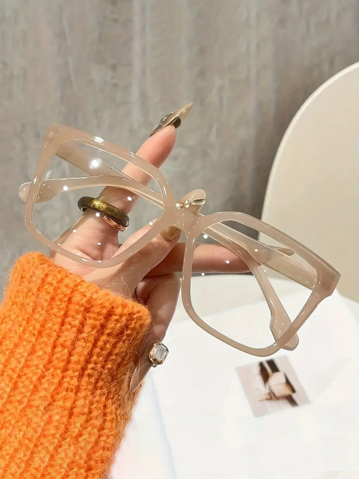 A hand holds a pair of elegant geometric reading glasses with transparent lenses against a light background.