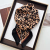 A luxury leopard print silk scarf tied in a bow, displayed in a brown box.