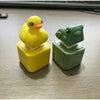 A yellow rubber duck fidget toy and a green frog fidget toy resting in their clear plastic cases on a wooden desk.