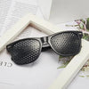 A pair of black vision correction pinhole glasses resting on a white background next to some paper.