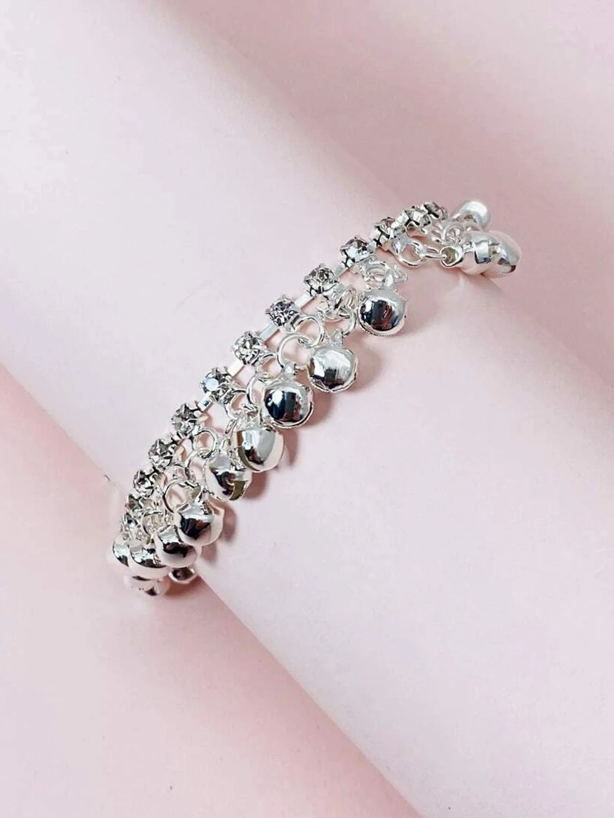 Silver bell foot chain anklet bracelet with small silver charms on a pink background.