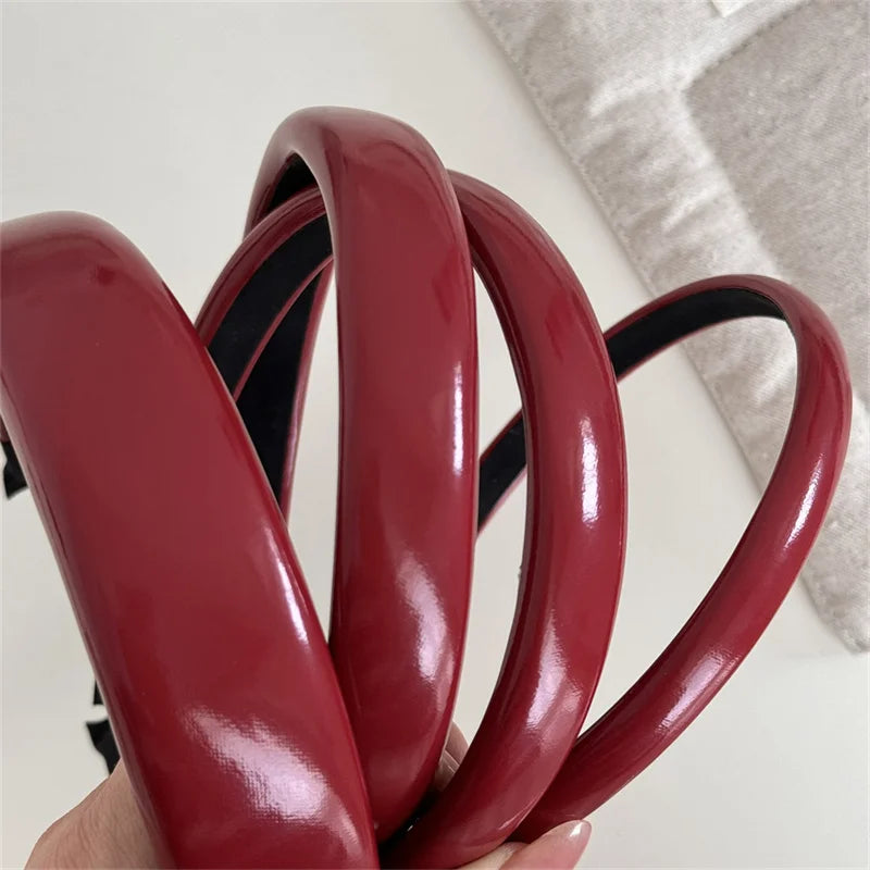 A hand holds several glossy maroon PU leather fashion headbands for women.