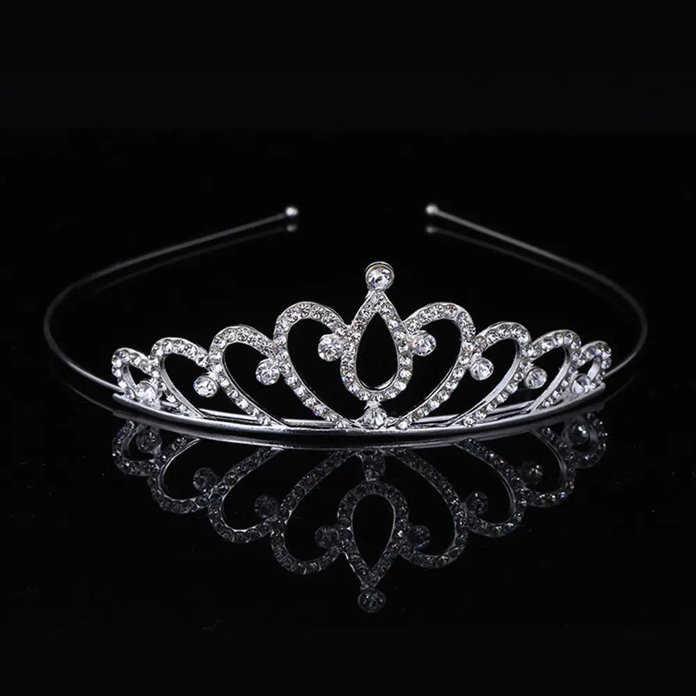 A silver crystal hair crown for women with a detailed royal design, displayed on a black reflective surface.