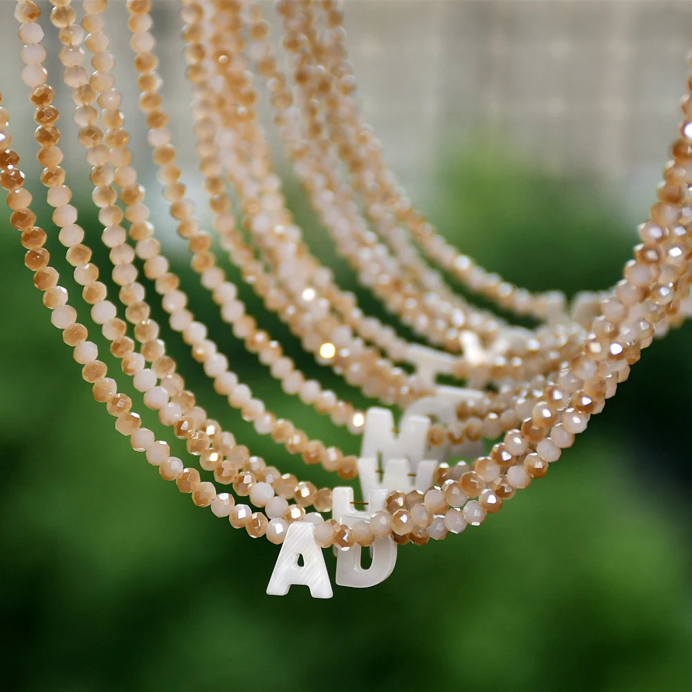 A close-up of a delicate necklace featuring mother of pearl beads and crystal accents, with a white initial charm.