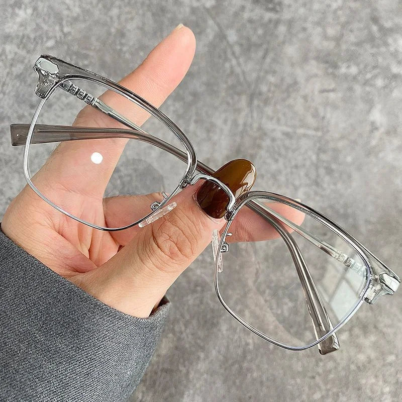 A hand holds a pair of clear metal half-frame reading glasses with brown tinted lenses against a gray background.