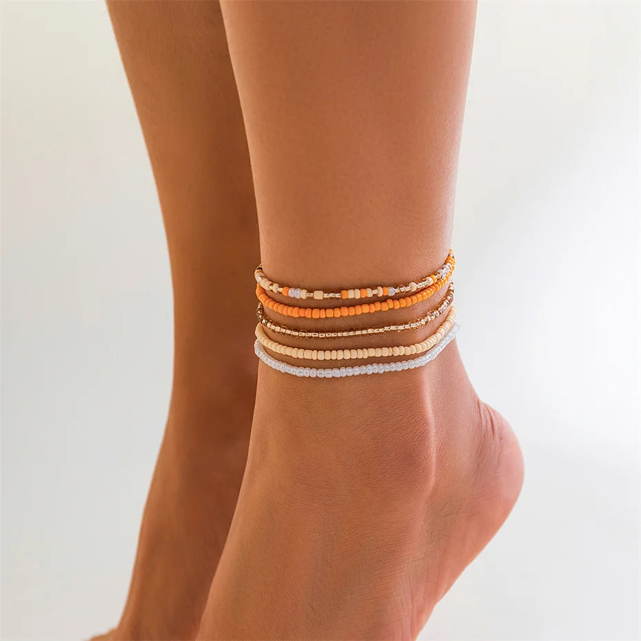 A close-up of a woman's ankle showing several stacked, colorful seed bead anklet bracelets.