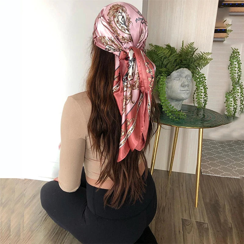 A woman with long brown hair is wearing a pink silk print shawl while sitting next to a green plant on a gold-legged table.