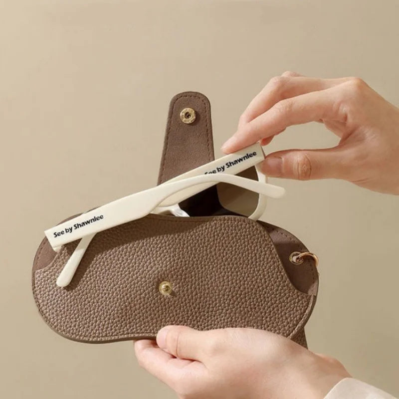 A hand holds a brown PU leather eyewear storage case with a white strap and a gold clasp.