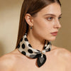 A woman models a silk scarf with a black and white polka dot print tied around her neck, featuring a pearl accent.