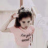 A young girl wearing a crystal hair crown and pink shirt looking down.