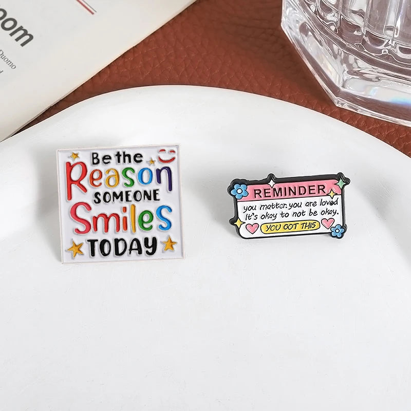 An enamel lapel pin with the text Be the Reason Someone Smiles Today featuring colorful stars and text on a white background.